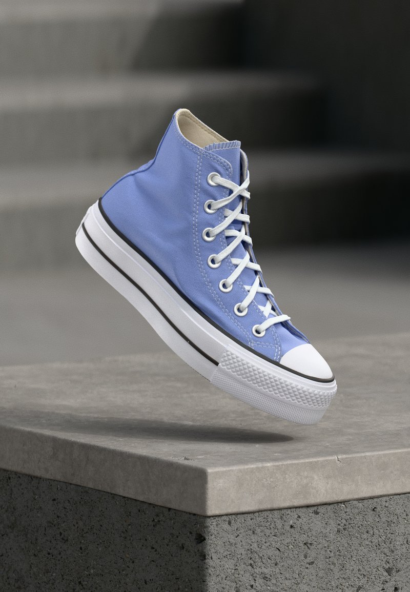 Μπλε καμβάς αθλητικό παπούτσι με λευκά κορδόνια και καουτσούκ σόλα που αιωρείται πάνω από μια τσιμεντένια άκρη, γκρι σκαλοπάτια θολωμένα στο φόντο.