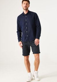 Camisa de botones azul marino con cuello y mangas largas, combinada con pantalones cortos enrollados azul marino y zapatillas blancas de caña alta.