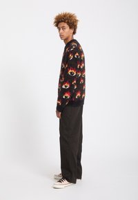 Jeune homme aux cheveux bouclés, debout de profil, portant un pull noir duveteux avec des motifs de flammes et d'yeux rouges et jaunes, pantalon noir et baskets claires.