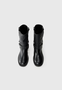 Botas de cuero negras hasta la mitad de la pantorrilla con un acabado texturizado, que cuentan con una correa con acentos metálicos y lengüetas en el caño para facilitar su uso.