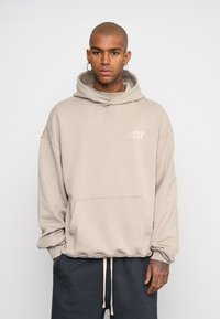 Felpa beige con tasca a marsupio, cappuccio con cordino e logo discreto sul petto. Il tessuto sembra morbido e strutturato.