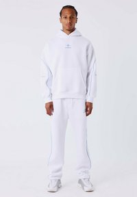 Jonge man met gevlochten haar, gekleed in een witte hoodie en joggingsbroek met blauwe accenten en witte sneakers, staand tegen een effen achtergrond.