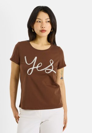 Mujer con cabello oscuro que lleva aretes de aro dorados, una camiseta marrón con la palabra "Sí" y pantalones blancos, de pie frente a un fondo liso.