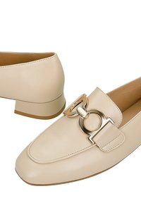 Beżowe skórzane mokasyny na niskim obcasie z kwadratowym noskiem, z dekoracyjną złotą okrągłą metalową klamrą na górze.