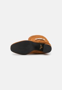 Botte mi-mollet en daim camel avec semelle en caoutchouc noire, détail boucle métallique et petit talon bloc, vue de dessous.