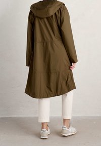 Olivgrön vattentät jacka med huva, åtdragen midja och sidofickor. I kombination med beige avslappnade byxor och vita sneakers.