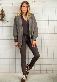 Grijze gebreide cardigan over een donkerbruin, aansluitend outfit. Getextureerde stof, lange mouwen en bruine sportieve schoenen met witte en beige accenten.