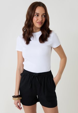 Kobieta o falowanych brązowych włosach nosi dopasowany biały t-shirt, czarne szorty ze sznurkiem i masywne bransoletki, stoi na jednolitym tle.