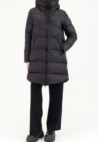 Zwarte pufferjas met een capuchon, met ritsluiting, horizontaal doorgestikt ontwerp en zijzakken. Gemaakt met zwarte broek en chunky zwarte schoenen.