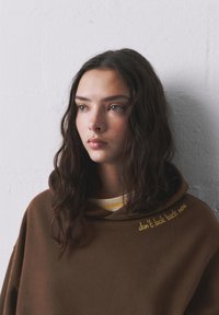 Hnedá mikina s kapucňou a žltou výšivkou s nápisom "nezapomínaj sa teraz pozerať späť." Má voľný strih a mäkký, textúrovaný materiál.