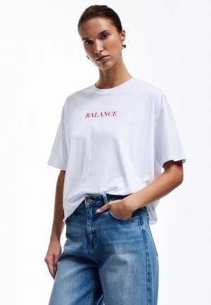 T-shirt en coton blanc avec le texte rouge "BALANCE", coupe décontractée, manches courtes. Assorti à un jean en denim bleu taille haute.