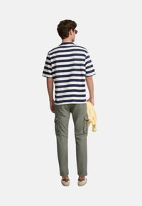 Camiseta de manga corta a rayas en navy y blanco, pantalones cargo oliva y un suéter amarillo claro colgado del brazo. Estilo casual con un corte relajado.