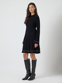 Mulher com cabelo castanho longo vestindo um vestido preto de manga longa com nervuras e detalhes brancos, e botas de couro pretas até ao joelho, de pé de lado.