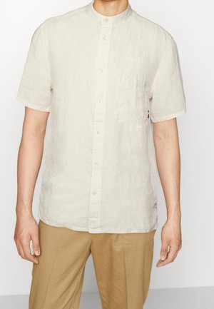 Homme portant une chemise en lin écru à manches courtes avec un col mandarin et un pantalon beige sur un fond uni.