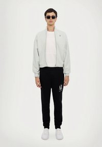 Ανοιχτό γκρι μπουφάν με φερμουάρ πάνω από λευκό t-shirt, συνδυασμένο με μαύρη φόρμα με λογότυπο και λευκά αθλητικά παπούτσια. Συμπληρώνεται με γυαλιά ηλίου.