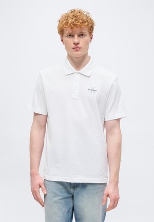 Νέος άνδρας με κατσαρά κόκκινα μαλλιά, που φοράει λευκό πόλο με κοντά μανίκια και ανοιχτό μπλε τζιν, στέκεται μπροστά σε απλό φωτεινό φόντο.