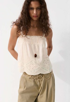 Mujer con cabello rizado que lleva una camiseta sin mangas color crema con encaje, pantalones beige, un collar largo con colgante y un gran cinturón de cadena dorada.