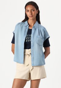 Lyseblå kortermet denimskjorte med knappelukking og brystlomme, brukt over en marineblå t-skjorte, sammen med beige shorts med knyttet midje.