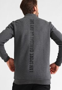 Suéter gris con cuello alto, que presenta un estampado de texto vertical negro en la parte posterior. Está hecho de una tela suave y texturizada con puños acanalados.