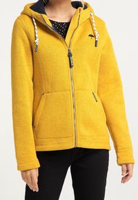 Gelbe Fleecejacke mit Reißverschluss, zwei Taschen, verstellbarer Kapuze und marineblauer Fütterung. Verfügt über weiße Kordeln und ein kleines Logo.