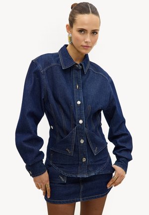 Femme portant une veste en jean bleu foncé ajustée avec des manches bouffantes et une mini-jupe en jean assortie, accessoirisée par de grandes boucles d'oreilles rectangulaires.