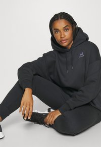 Felpa nera con cappuccio e cordino, tasca frontale e dettaglio del logo. Abbinata a pantaloni sportivi neri e scarpe da ginnastica nere. Tessuto liscio.
