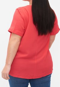 Blouse corail à manches courtes avec une texture douce et un ourlet arrondi, associée à un jean en denim bleu. Présente un détail discret de couture au dos.