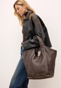 Brašna iz rjave usnjene tote z teksturirano površino, velika odprta zgornja stran in kratki ročaji. Na sprednji strani je poudarek z zlatim logotipom.
