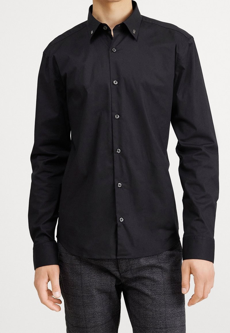 Mann trägt ein eng anliegendes schwarzes Hemd mit langen Ärmeln und dunkelgraue karierte Hose vor weißem Hintergrund.