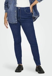 Tmavě modré skinny džíny z denimu s mírně zvýšeným pasem, minimálními švy a předními kapsami, v kombinaci s volnou denimovou košilí.