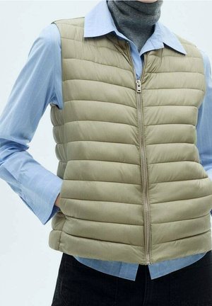 Personne portant un gilet matelassé beige zippé sur une chemise bleue boutonnée avec un col roulé gris en dessous.
