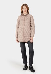 Beige gewatteerde jas met een gestructureerde afwerking, knopen sluiting aan de voorkant en een fuzzy kraag, gecombineerd met donkere jeans en zwarte chunky laarzen.