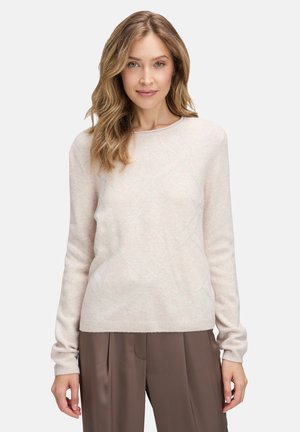 Hellbeige Pullover aus weichem Material, mit einem dezenten Rautenmuster und gerippten Bündchen, kombiniert mit hochgeschnittenen braunen Hosen.