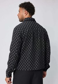 Un homme vu de dos, portant une veste noire avec un motif de chaîne blanche répétée et un pantalon noir, sur un fond uni.