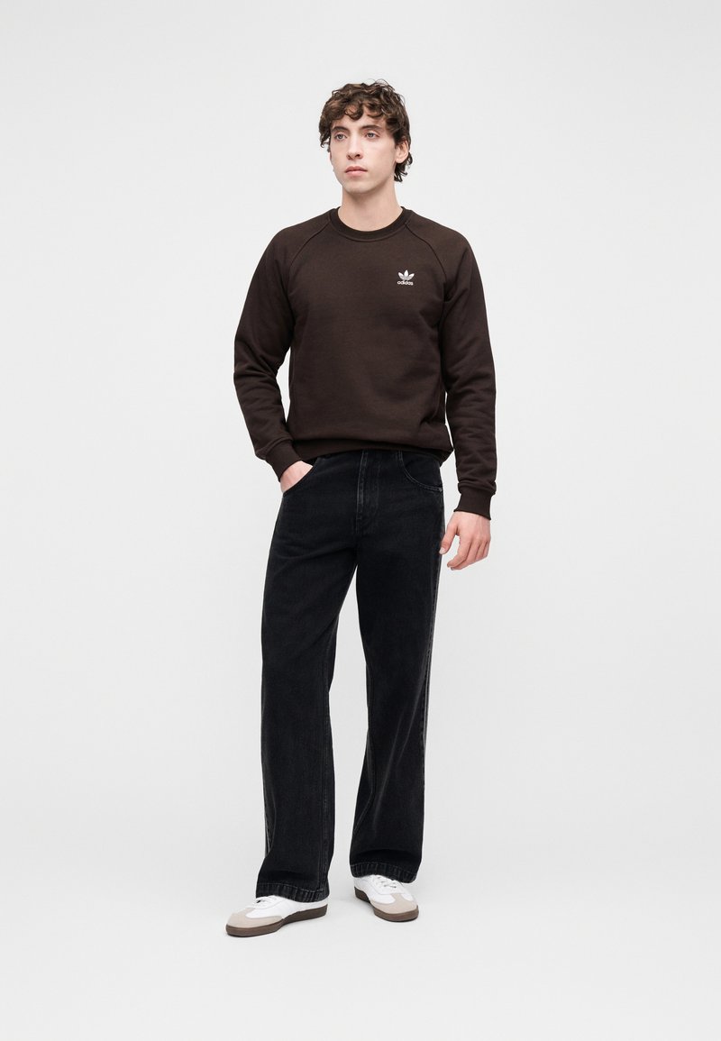 Brun sweatshirt med ribbad krage och broderad logotyp, kombinerad med löst sittande svarta jeans och vita sneakers med bruna detaljer.