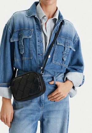 Mujer vestida con mono de mezclilla azul y camiseta de rayas, llevando un pequeño bolso cruzado negro tejido.