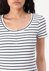 Ribbed, lühikeste varrukatega valge topp tumedate mereväe horisontaalsete triipudega. Omab ümmargust kaela ja kolme nupuga sulgemist ees.