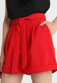 Red high-waisted shorts with a fabric belt, side pockets, and rolled hems. Made from smooth, lightweight material.