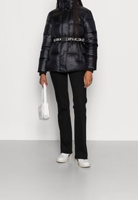 Osoba v černé prošívané bundě s páskem Calvin Klein, černých kalhotách, bílých teniskách a s bílou kabelkou na pozadí bez vzoru.