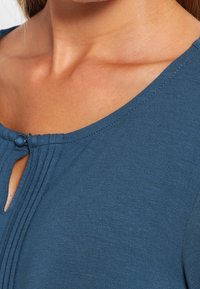 Tyrkysový top s kulatým výstřihem, se malým knoflíkem a detaily ve tvaru klíčové dírky, vyrobený z měkké tkaniny s texturovaným povrchem.