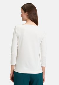 Haut à manches longues blanc avec un col carré, design ajusté, texture lisse et un léger éclat, associé à un pantalon teal.