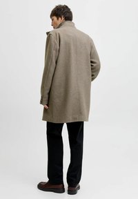 Oliivroheline villasegu mantli, millel on kõrge krae, nööpidega mansetid ja sirge lõige. Kombineeritud mustade pükste ja pruuni nahast kingadega.