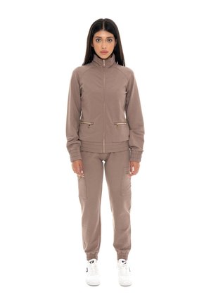 Le modèle porte un survêtement taupe zippé avec des poches cargo et des baskets blanches. Il se tient devant un fond blanc uni.