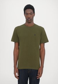 Tricou din bumbac de culoare verde măslin, cu mâneci scurte, decolteu rotund și un mic emblemă neagră pe piept. Purta cu blugi negri din denim.