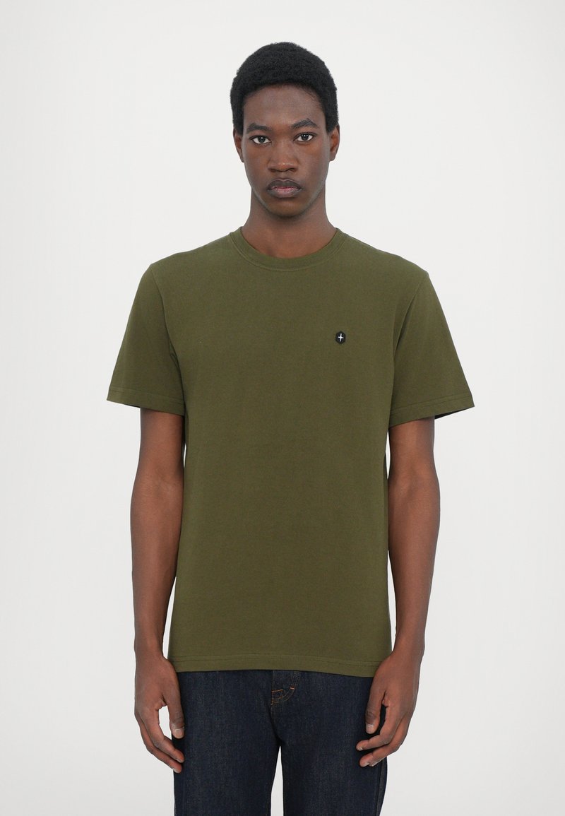 Tricou din bumbac de culoare verde măslin, cu mâneci scurte, decolteu rotund și un mic emblemă neagră pe piept. Purta cu blugi negri din denim.