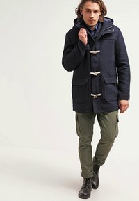 Abrigo de duffle azul marino con cierres de botón, bolsillos frontales y capucha, combinado con pantalones cargo verdes oliva y botas negras con cordones.