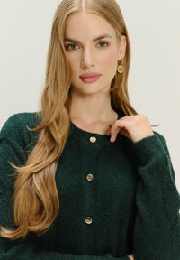 Blondynka z długimi włosami, ubrana w ciemnozielony sweter z guzikami i złote okrągłe kolczyki, dotykająca kołnierza, na neutralnym tle.