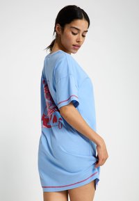 Zila kokvilnas T-kreklu kleita ar brīvu piegriezumu, īsām piedurknēm un sarkanu grafisko apdruku aizmugurē, ar kontrastējošām dūriens detaļām.