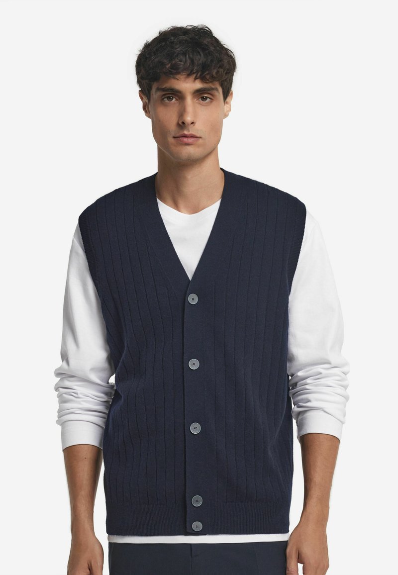Marinblå stickad väst med vertikal ribbing, V-ringning och fem knappar fram. Bärs över en vit skjorta och har en figurnära design.