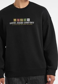 Czarna bluza z pięcioma kolorowymi kwadratowymi logo nad napisem "GUESS JEANS COMPANY" oraz nazwami miast Los Angeles, Nowy Jork, Londyn, Amsterdam.